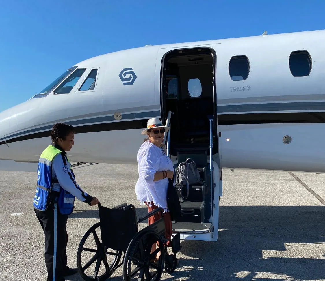 A woman in a white sun hat, sunglasses and white blouse stands at the steps of a small white jet on a sunny tarmac, beside an airport worker in a safety vest holding a wheelchair.