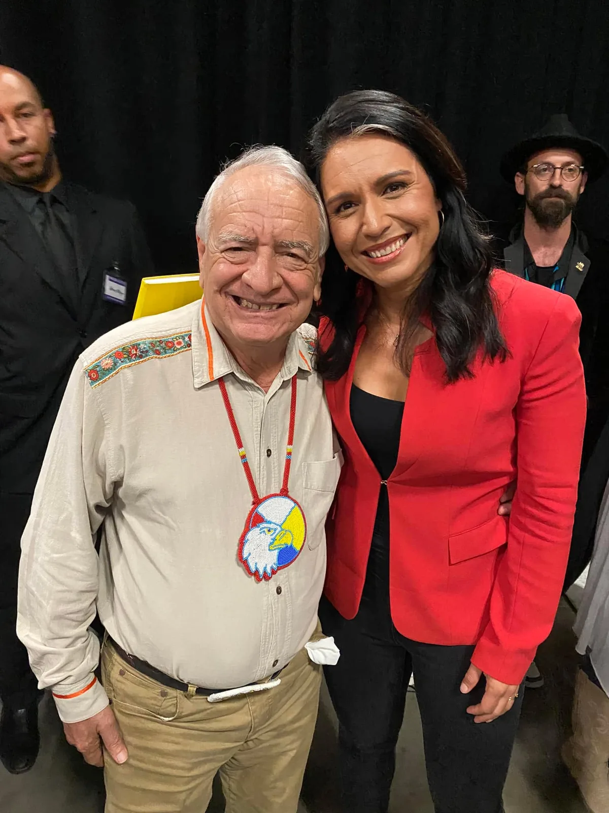 Phil Lane Jr., an elder with white hair wearing a beaded eagle medallion and embroidered shirt, smiles beside Tulsi Gabbard in a red blazer at an indoor event.
