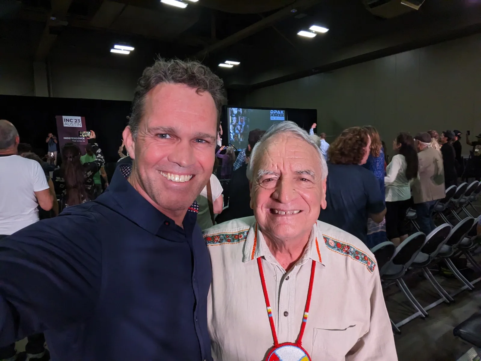 A smiling man in a dark shirt takes a selfie with Phil Lane Jr., an elder in an embroidered shirt and beaded medallion, at a crowded INC'23 Austin conference hall.