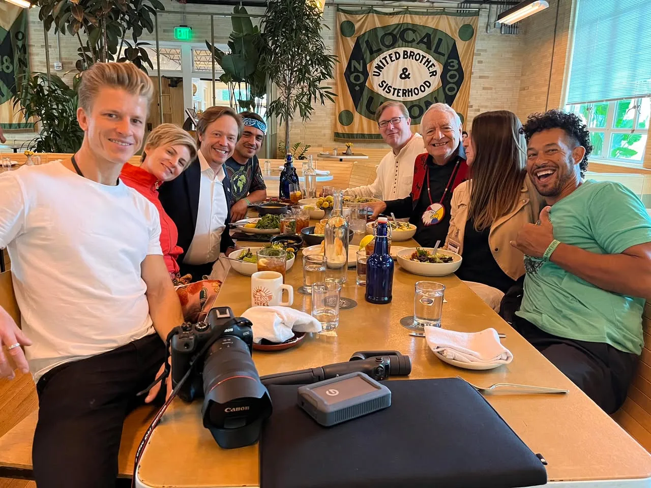 Eight people smile around a restaurant table with meals, drinks and a camera, beneath a banner reading "United Brother & Sisterhood." An elder in a red vest and eagle medallion sits among them.