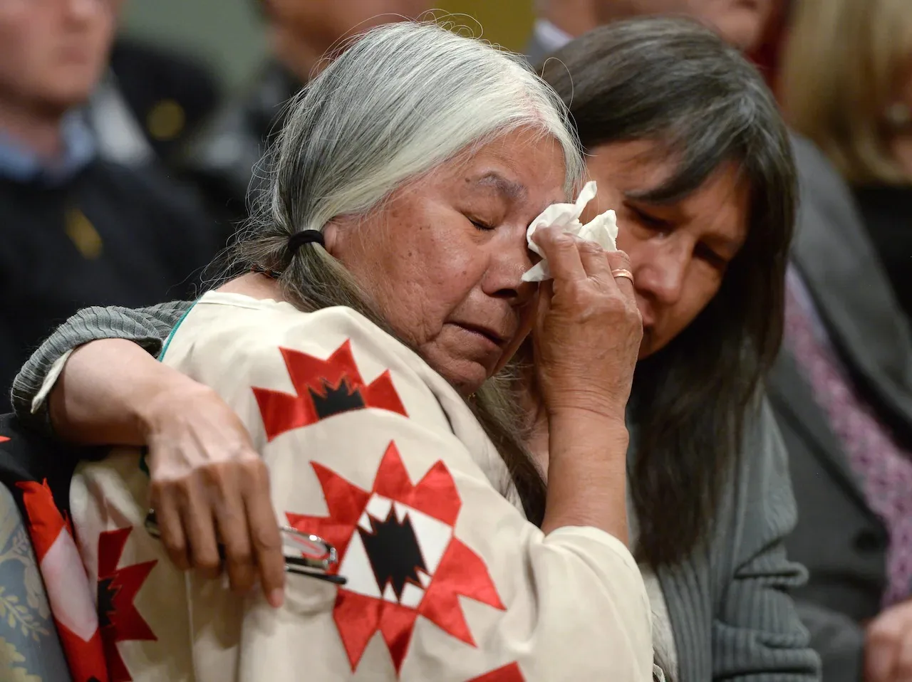 An elder woman with long grey hair, wearing a cream shawl with red and black star designs, wipes tears with a tissue as another woman embraces and comforts her.