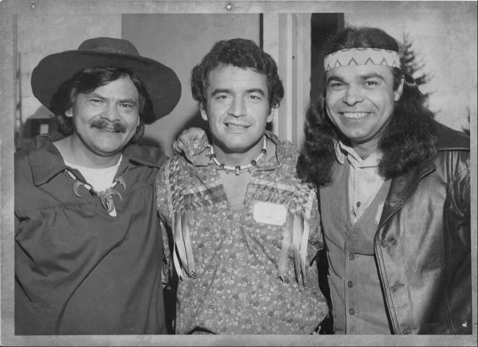 Black-and-white 1975 photo of Phil Lane Jr., Gary Hallaire and Phil Lucas smiling arm in arm, wearing a wide-brimmed hat, a ribbon shirt and a beaded headband.