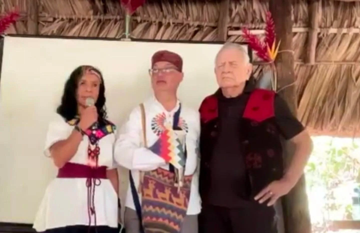 Three people stand beneath a thatched roof decorated with red flowers: a woman in a white embroidered blouse speaking into a microphone, a man in a white shirt and woven bag, and an older man in a black and red vest.