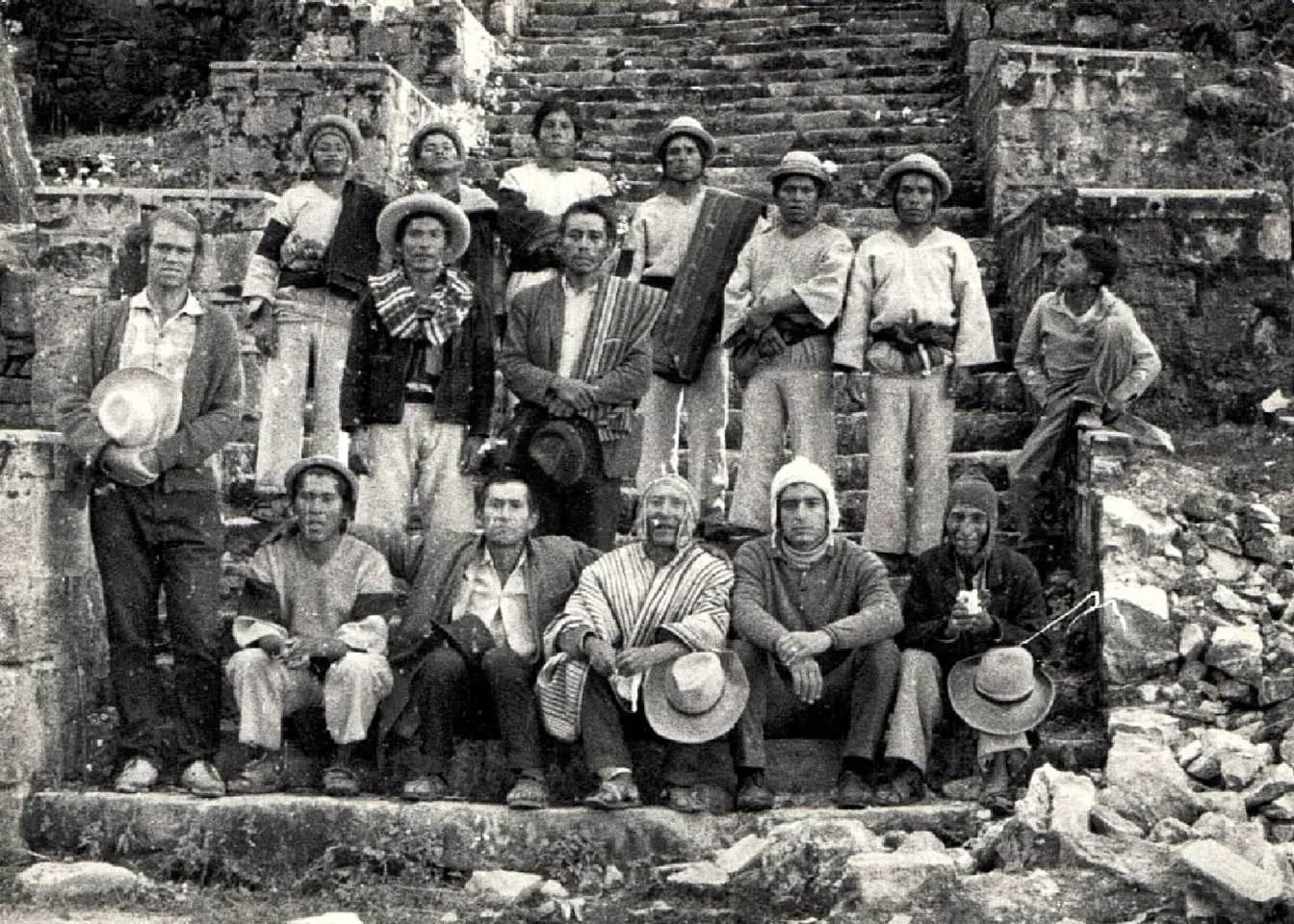 Black-and-white photograph of a group of Quechua men in hats, ponchos and woven cloth posed on ancient stone stairs, some seated, with a young non-Indigenous man standing at left.