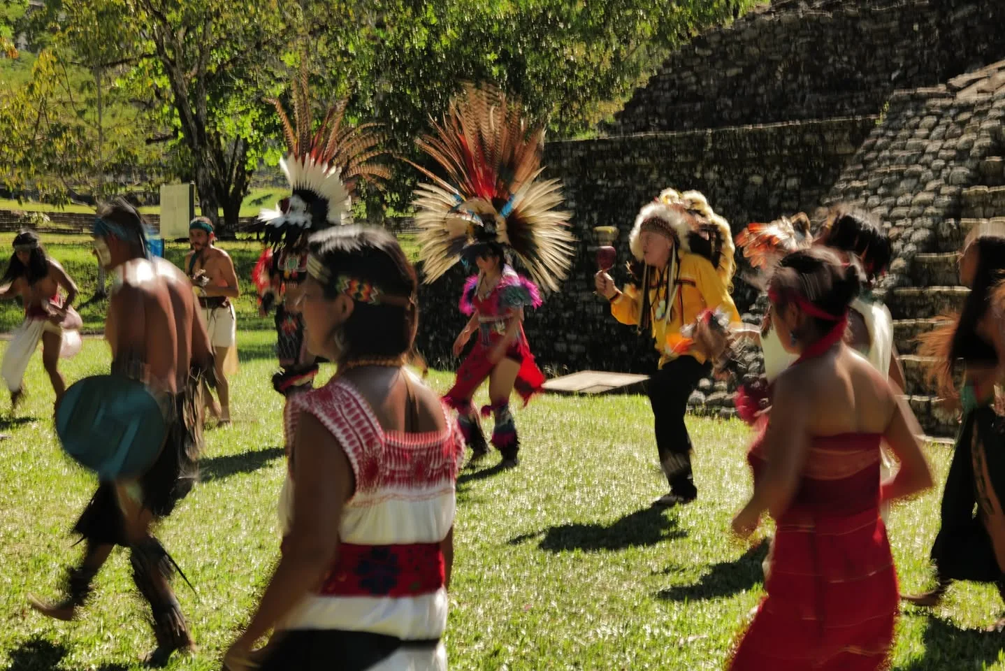 Dancers in traditional dress and feathered headdresses move across a sunlit lawn beside an ancient stone pyramid; an elder in a yellow shirt and feather headdress shakes a rattle.