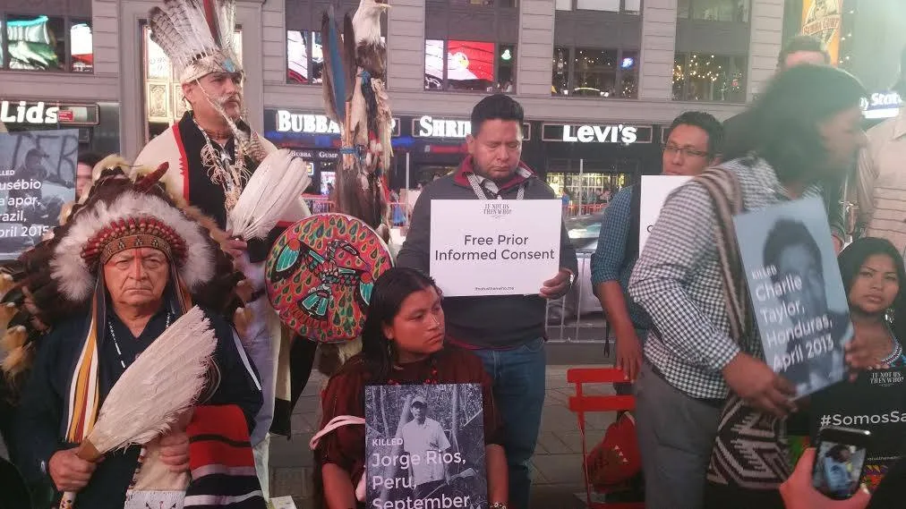 Indigenous people gather at night in Times Square, elders in feather headdresses holding eagle feathers and a painted drum, others holding signs for Free Prior Informed Consent and memorials to killed land defenders.