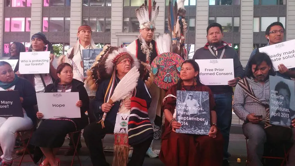 Indigenous leaders sit and stand in Times Square holding signs reading Hope, Who is protecting our forests?, and Free Prior Informed Consent, plus portraits of defenders killed in Peru, Brazil and Honduras.