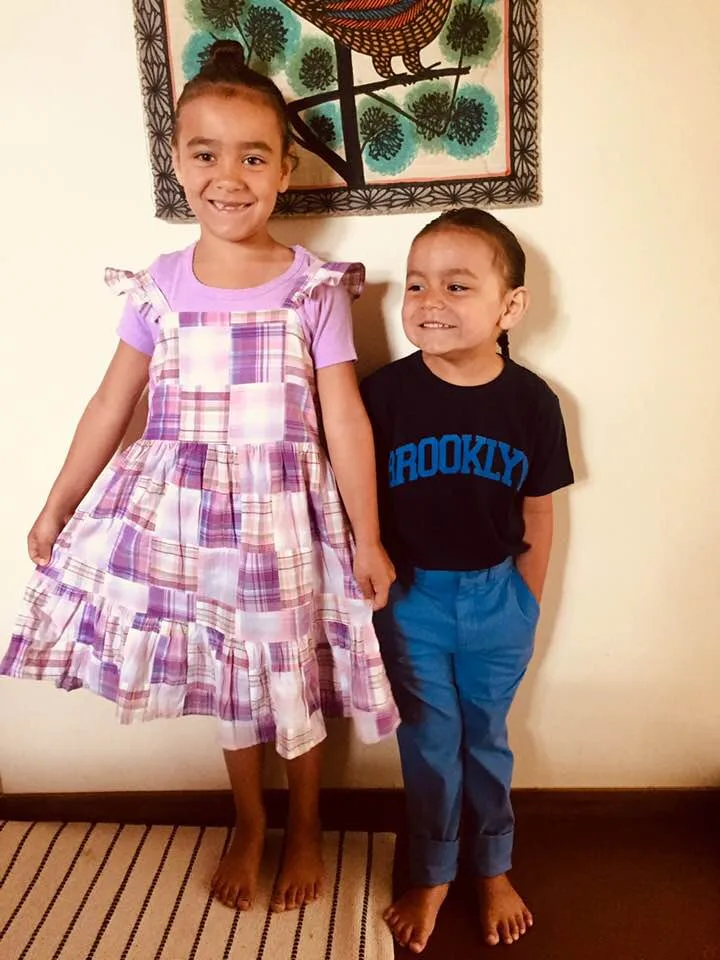 Two smiling barefoot children stand against a wall beneath a bird tapestry, a girl in a purple patchwork dress and a boy in a Brooklyn T-shirt and blue pants.