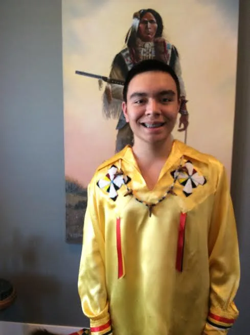A smiling young man in a yellow satin ribbon shirt with beaded rosettes stands before a painting of a Native warrior holding a rifle.