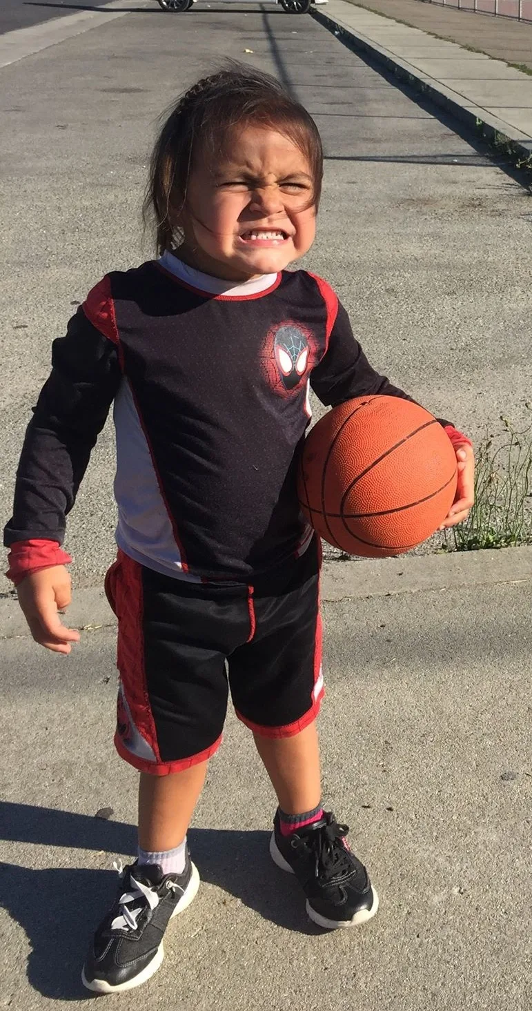 A young child in a black and red Spider-Man outfit stands on a sunny pavement holding a basketball, scrunching up their face in a fierce grimace.