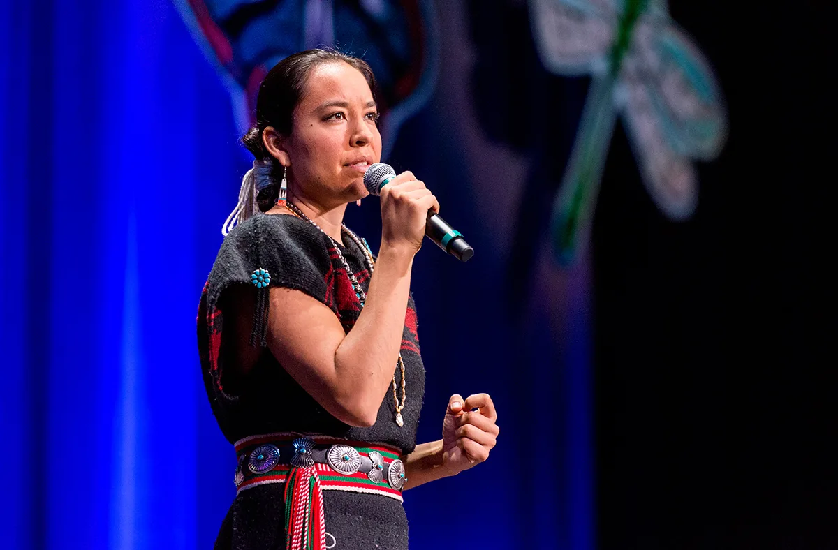 A young Indigenous woman speaks into a microphone on stage, wearing a black woven dress, a red and green sash, a silver concho belt and turquoise jewelry, before a blue backdrop.