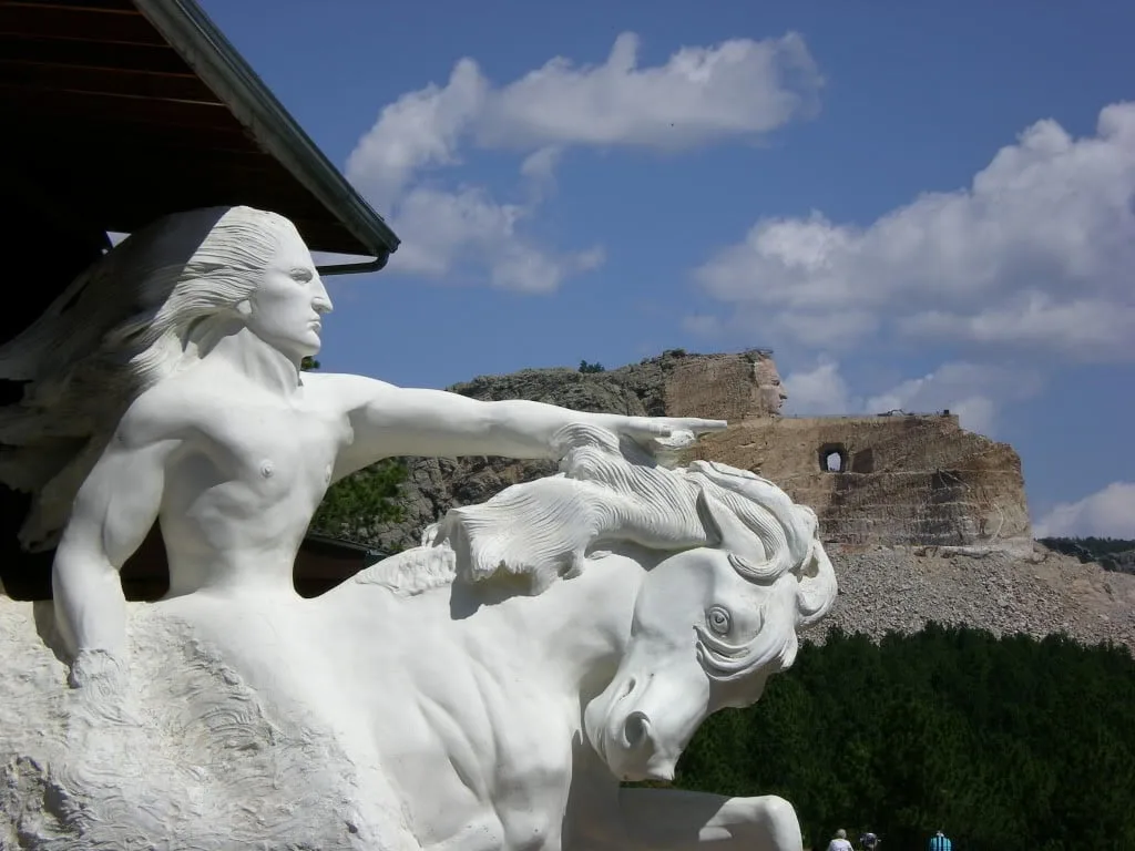 White sculpture of Crazy Horse on horseback pointing outward, with the partially carved Crazy Horse Memorial mountain under a blue sky behind it.
