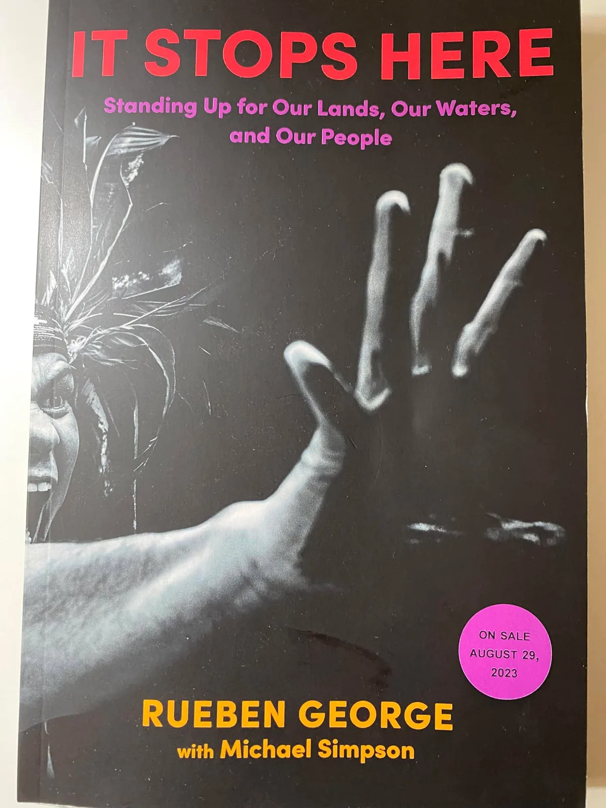 Cover of the book "It Stops Here: Standing Up for Our Lands, Our Waters, and Our People" by Rueben George with Michael Simpson, showing a black-and-white image of a man in a feathered headdress shouting with his hand raised. A sticker reads "On sale August 29, 2023."