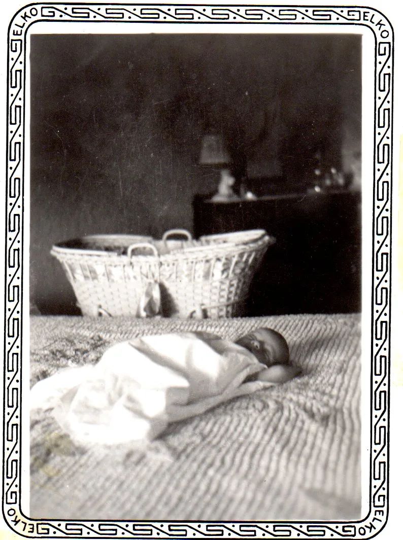 Old black-and-white snapshot with an ornate border marked "ELKO" of a newborn baby sleeping on a chenille bedspread, a woven basket and dresser lamp behind.