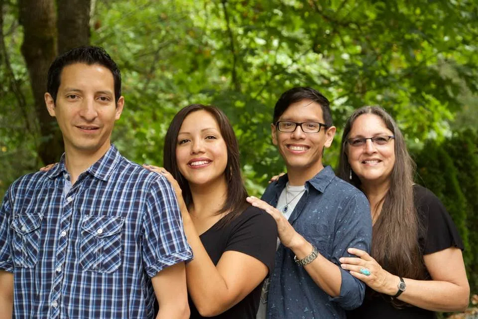 Four smiling relatives pose close together outdoors in front of green trees: two young men, a young woman, and an older woman with long hair and glasses, hands resting on each other's shoulders.