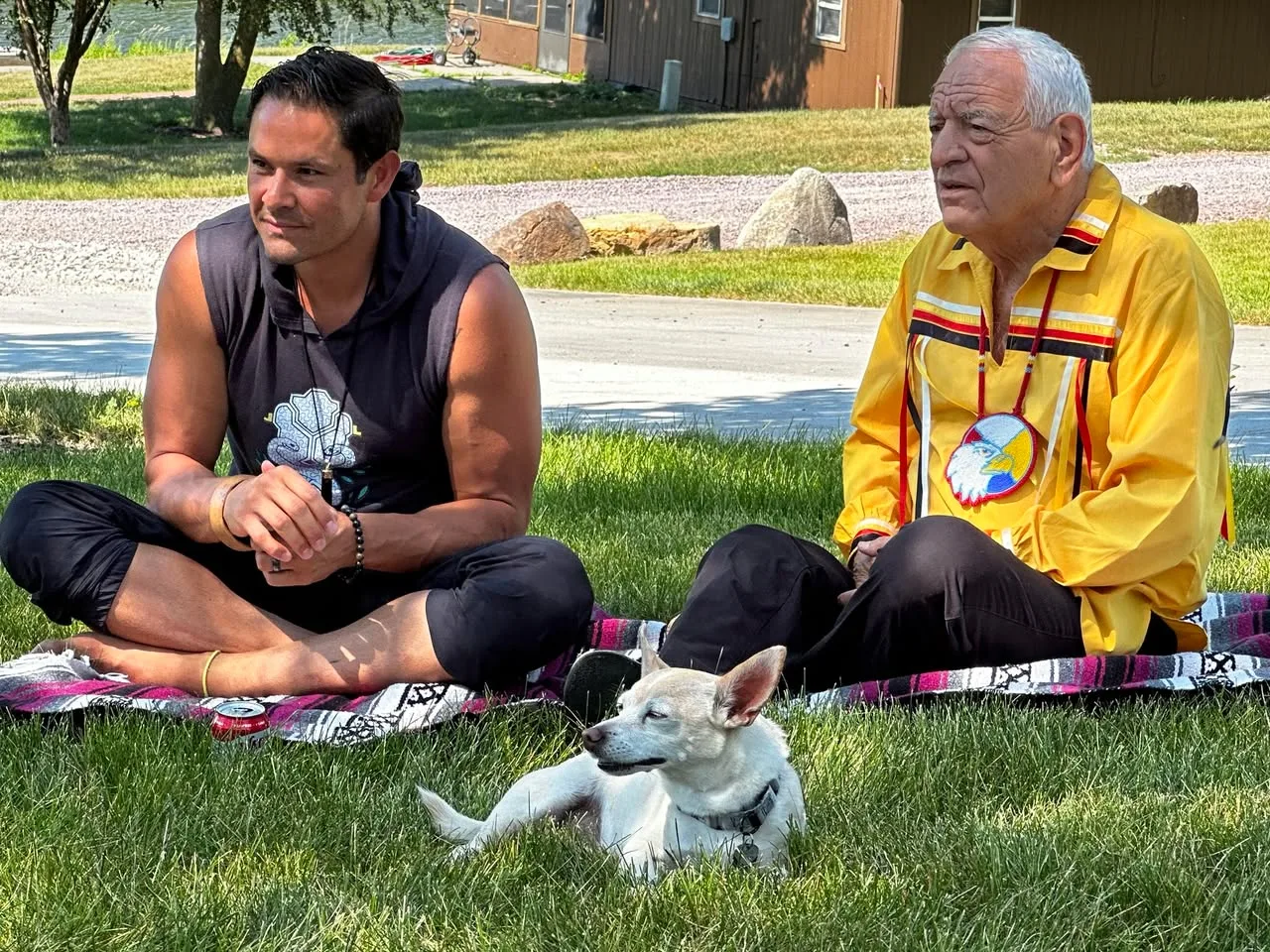 A younger man in a dark sleeveless hoodie and an elder in a yellow ribbon shirt with an eagle medallion sit cross-legged on a blanket on the grass, a small white dog resting in front.