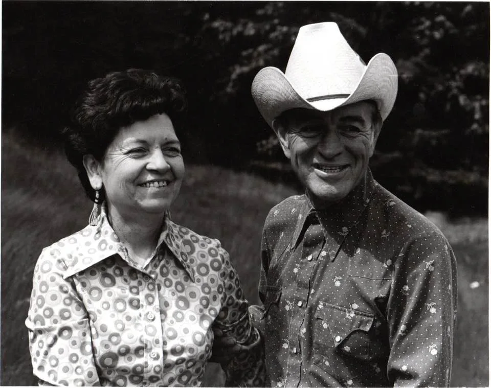 Black-and-white portrait of a smiling couple outdoors: a woman with short dark hair, earrings and a patterned blouse beside a man in a white cowboy hat and western shirt.