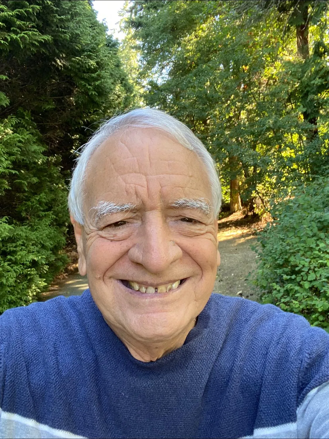 Selfie of a smiling elder man with white hair, wearing a blue sweater, standing on a sunlit path lined with tall green trees and shrubs.
