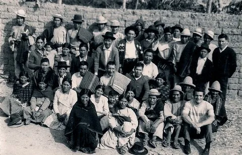 Black-and-white group photograph of several dozen Quechua women, men and children in hats and traditional dress, seated and standing before a stone wall in the Bolivian Andes.