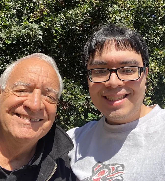Smiling selfie of an older white-haired man in a dark jacket beside a young man with black hair and glasses in a gray shirt with a Northwest Coast design, in front of green trees.