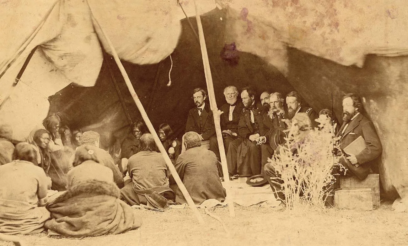 Sepia historical photograph of a treaty council inside a large tipi: bearded men in dark 19th-century suits and military uniforms sit facing Native American men and women seated on the ground.