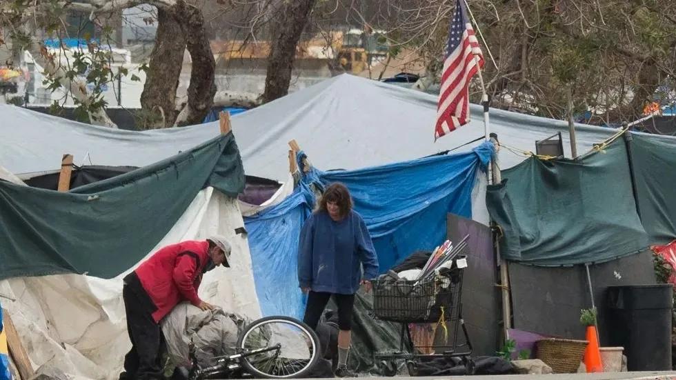A homeless encampment of tents covered in green, blue and white tarps under an American flag, with a man bent over a bicycle and a woman standing by a loaded shopping cart.