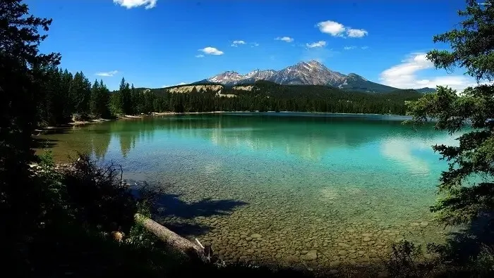 A clear turquoise lake with visible stones beneath the shallow water, surrounded by evergreen forest, with a snow-dusted mountain peak under a bright blue sky.