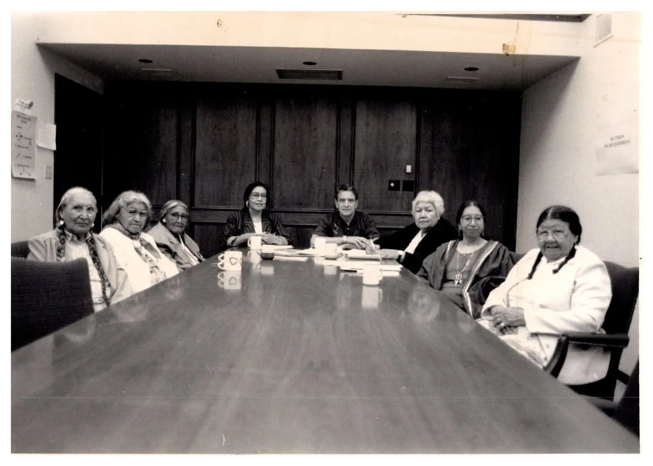 Black-and-white photo of eight people seated around a long conference table with coffee mugs, mostly grandmothers, some with long braids, and one man at the center.