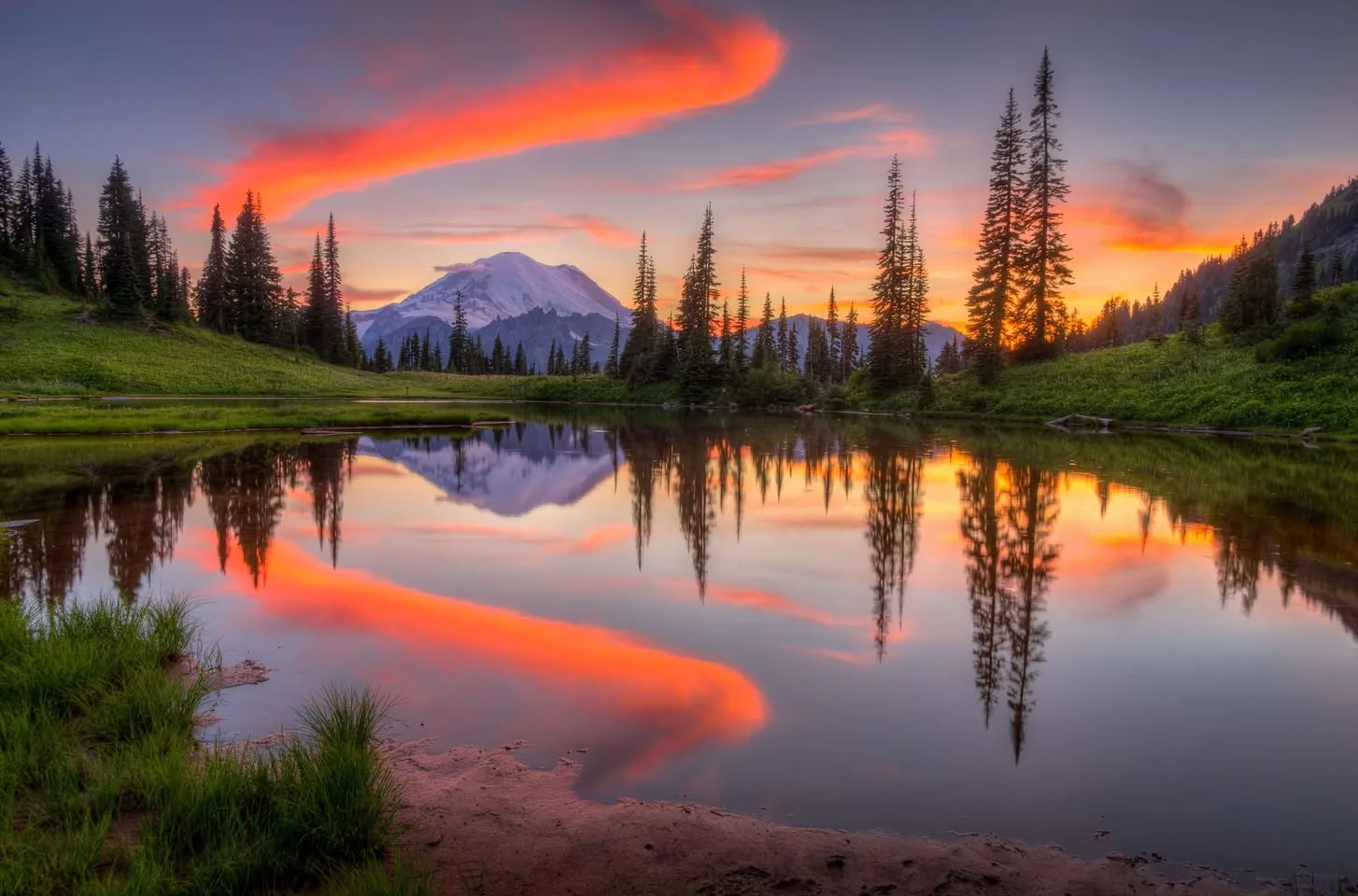 A snow-capped mountain at sunset framed by tall evergreens, with fiery orange clouds and the peak mirrored in a still alpine lake bordered by green grass.