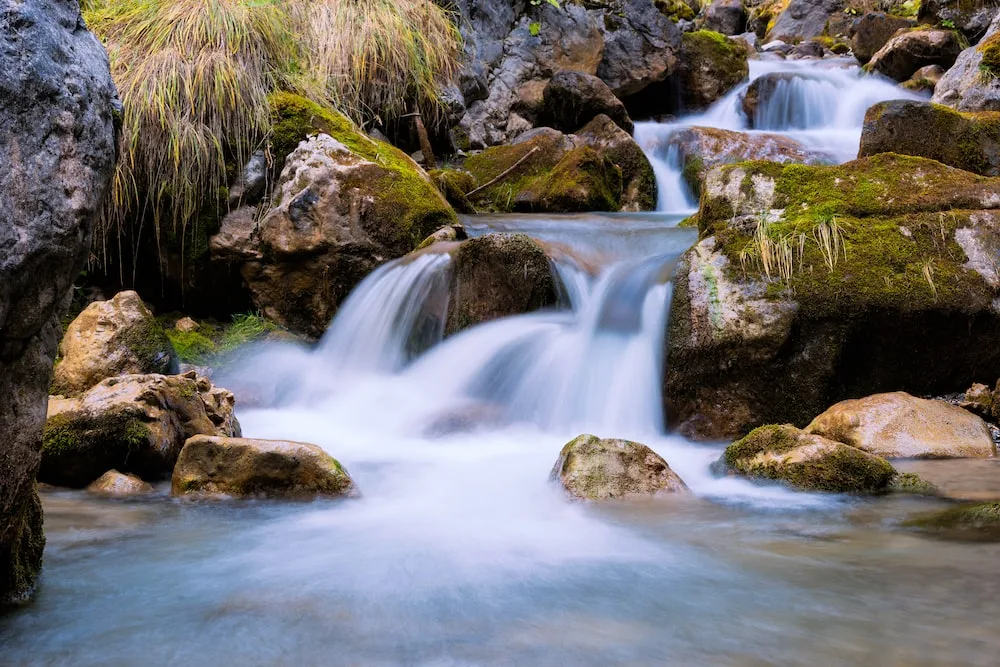 A mountain stream cascades in silky white falls over moss-covered boulders, with tufts of grass growing among the rocks.
