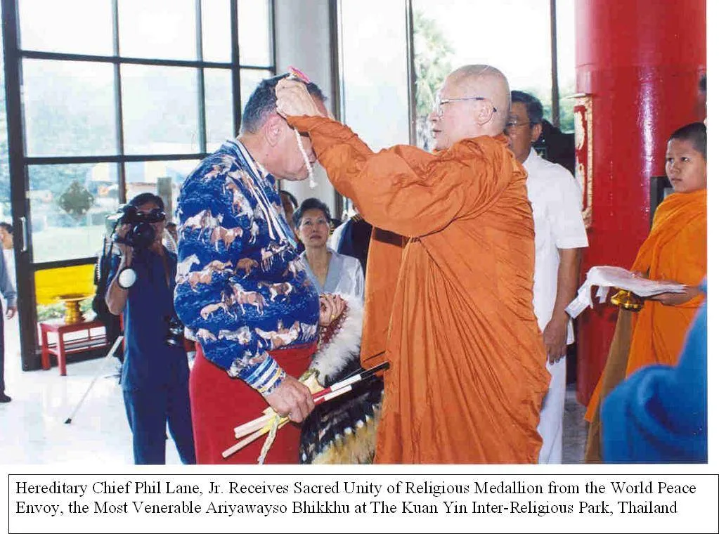 A Buddhist monk in saffron robes places a medallion on a cord around the neck of Hereditary Chief Phil Lane Jr., who wears a horse-print shirt and holds an eagle feather fan. A caption says he receives the Sacred Unity of Religious Medallion at the Kuan Yin Inter-Religious Park, Thailand.