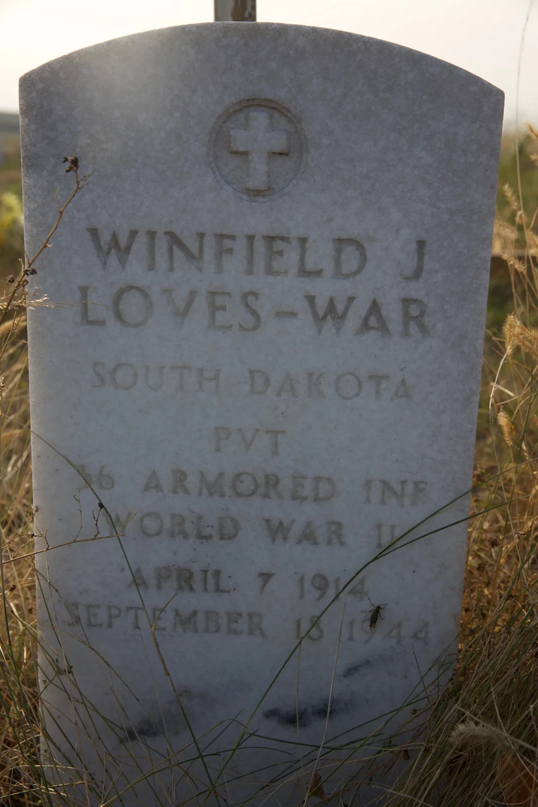 A weathered white military gravestone in dry prairie grass reading Winfield J Loves-War, South Dakota, PVT, Armored Inf, World War II, April 7 1914 – September 18 1944.