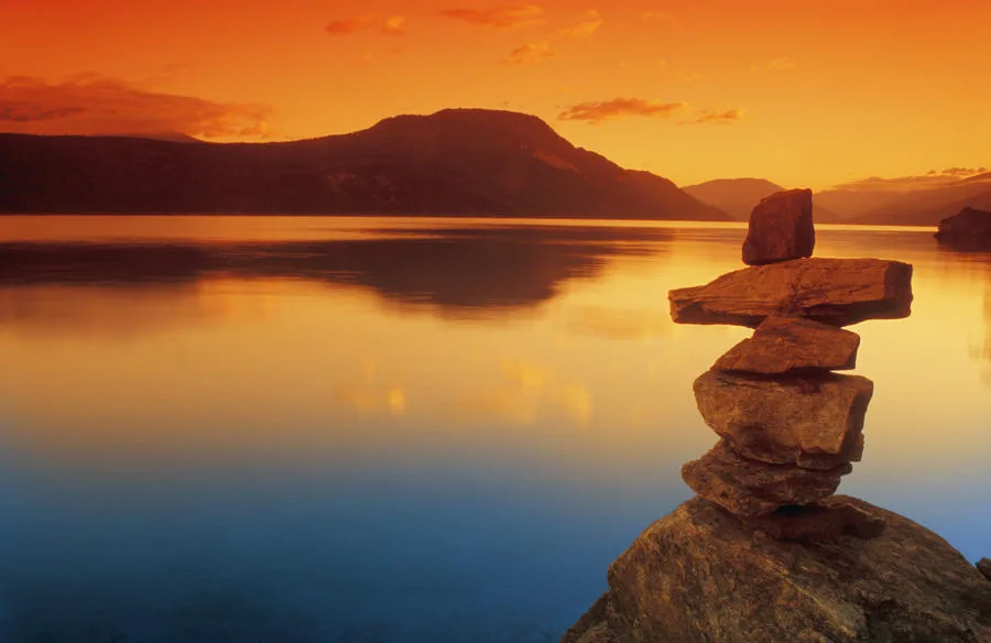 A stacked stone inukshuk stands on a rock beside a calm mountain lake, with hills and water glowing orange under a sunset sky.