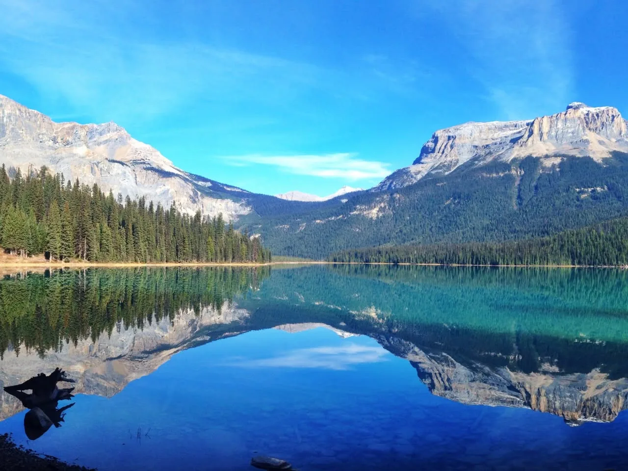 A calm turquoise mountain lake reflecting snow-dusted rocky peaks, evergreen forests and a bright blue sky, with driftwood at the shore.