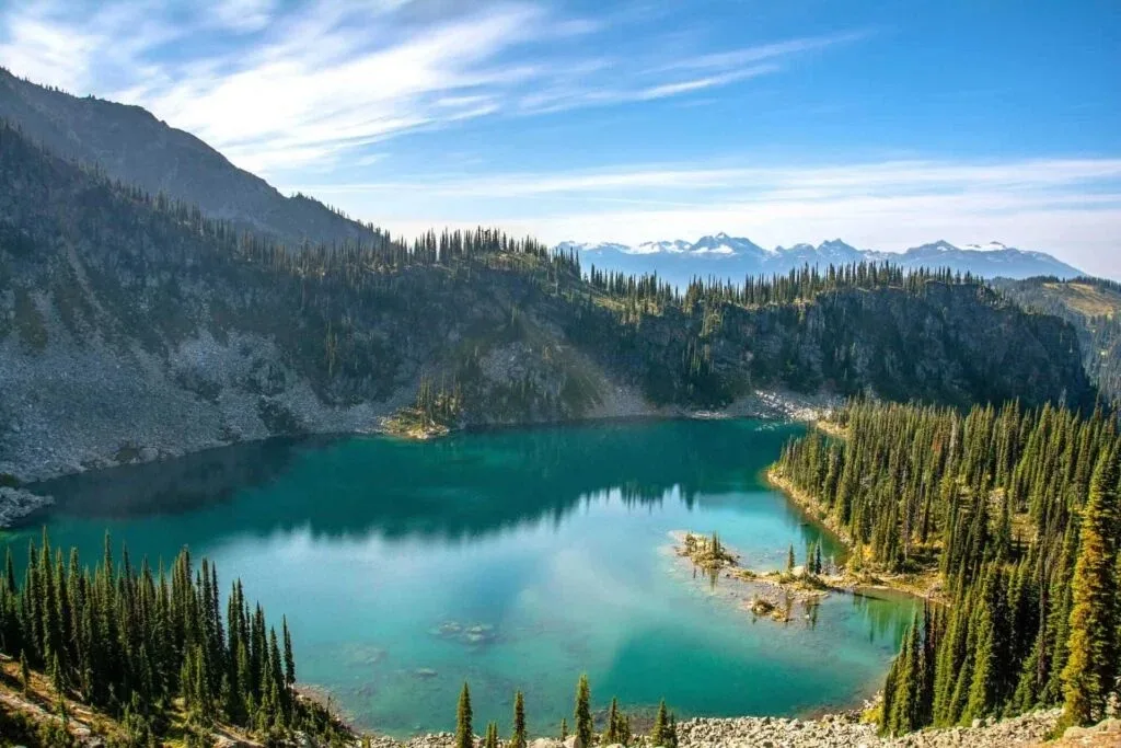 A turquoise mountain lake surrounded by evergreen forest and rocky slopes, with snow-capped peaks in the distance under a blue sky with wispy clouds.