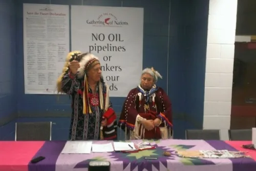 Chief Phil Lane Jr., in a feather headdress and regalia, raises an eagle feather fan beside Faith Spotted Eagle in a beaded dress, under a Gathering of Nations sign reading "NO OIL pipelines."