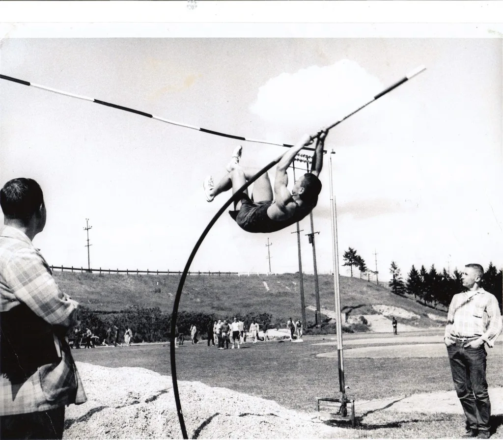 Black-and-white photo of a young pole vaulter arching over the crossbar on a bending fiberglass pole, as two men in plaid shirts watch beside a sand pit on a sports field.