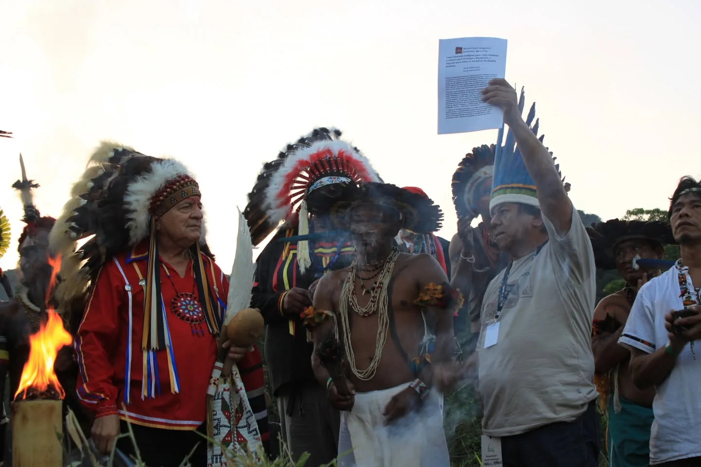 Indigenous leaders in feathered headdresses gather outdoors beside a small fire as smoke rises; one man holds a printed document aloft while another performs a smudging ceremony.