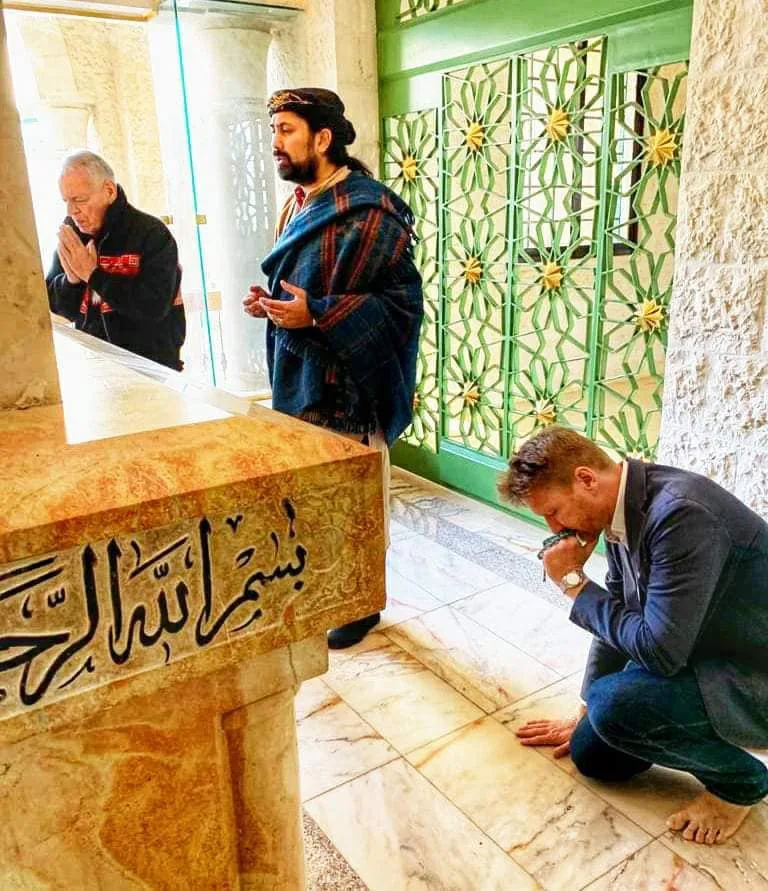 Three men pray inside a mosque beside a marble tomb inscribed with Arabic calligraphy: an older man with palms together, a man in a striped shawl, and a man kneeling barefoot.