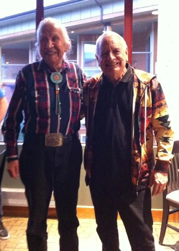 Two smiling white-haired elders stand side by side indoors, one in a striped shirt with a turquoise bolo tie and large belt buckle, the other in a colorful patterned jacket.