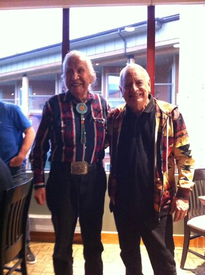 Two smiling elders stand side by side indoors. One has long white hair, a patterned shirt, beaded bolo tie and large belt buckle; the other wears a bright geometric-print jacket.