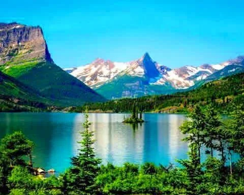A turquoise mountain lake with a small tree-covered island, framed by evergreens, beneath a sharp snow-dusted peak and a steep green cliff under a clear blue sky.