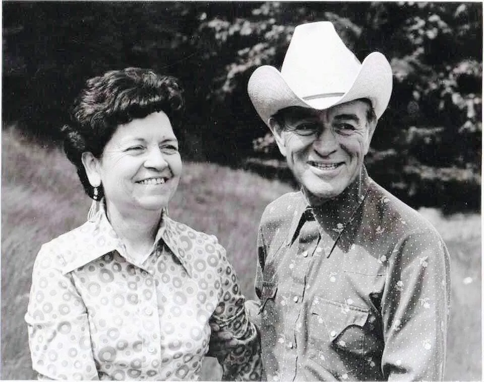 Black-and-white photo of Phil Lane Jr.'s mother and father standing close together outdoors, smiling; she wears a patterned blouse and earrings, he wears a white cowboy hat and western shirt.