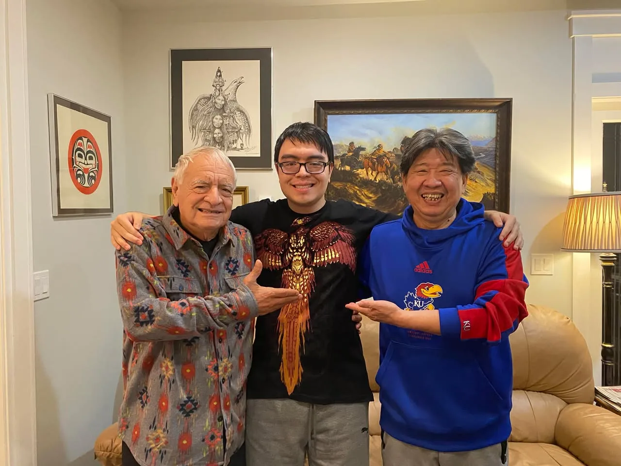 Three men stand arm in arm in a living room hung with Indigenous art: an older man in a patterned shirt, a young man in glasses and an eagle T-shirt, and a smiling man in a blue KU hoodie.