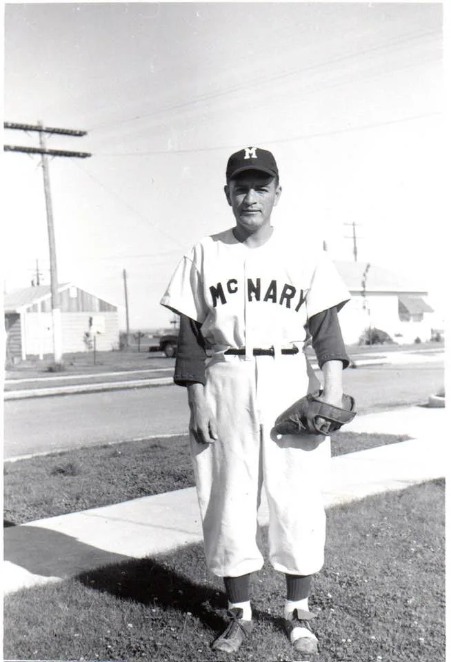 Black-and-white photo of a young man in a McNary baseball uniform, cap with an M, and fielder's glove, standing on a lawn beside a street with houses and power poles.