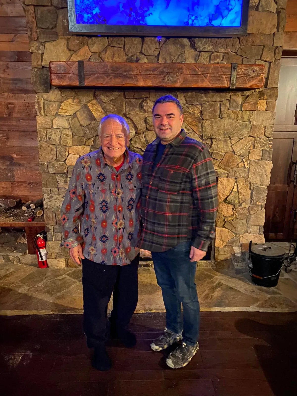 An elder in a patterned shirt with Indigenous-style designs stands smiling beside a younger man in a plaid flannel shirt, in front of a stone fireplace with a wooden mantel.