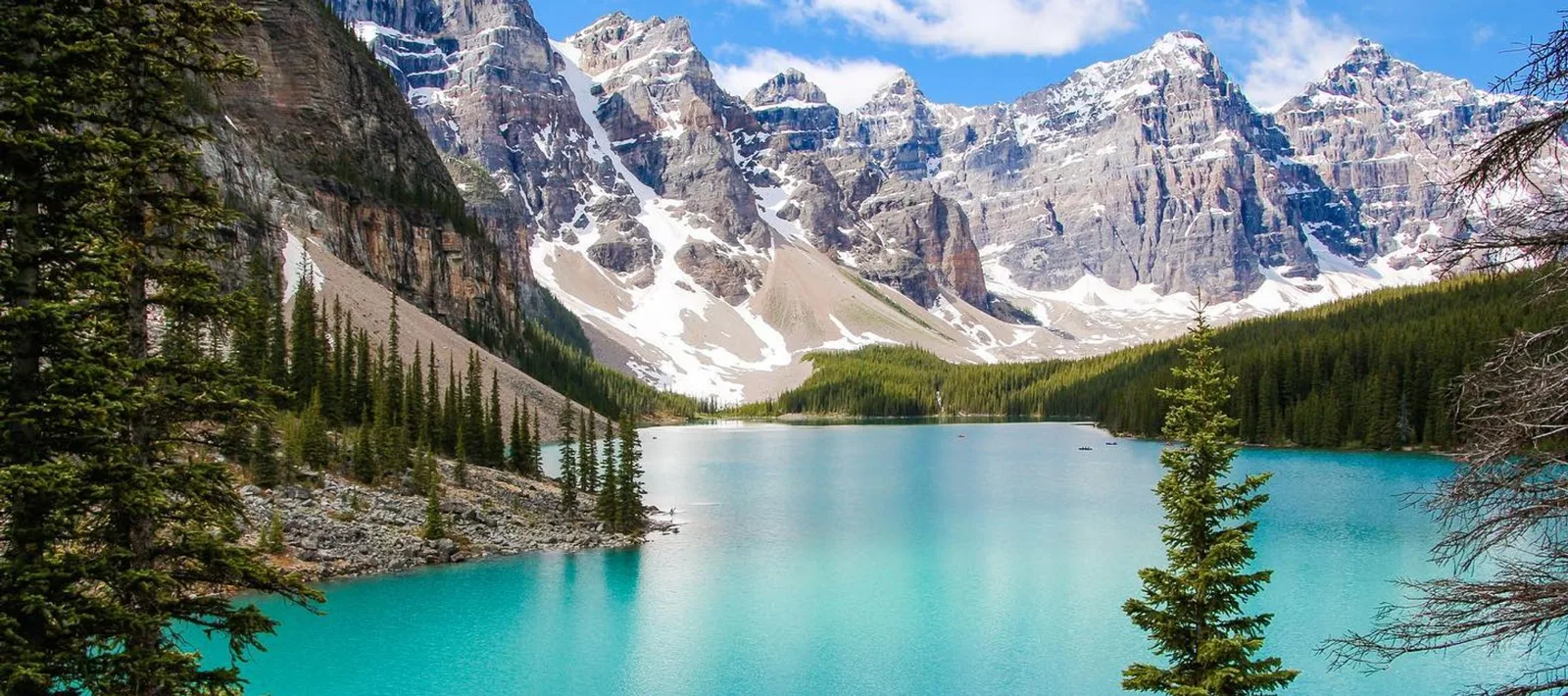 A turquoise mountain lake framed by tall evergreen trees, with rugged snow-streaked peaks rising beneath a blue sky with scattered clouds.