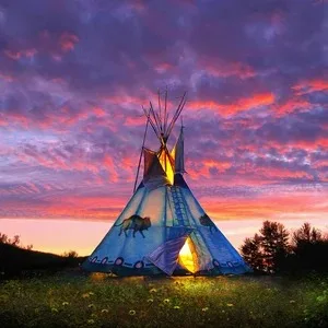 A tipi painted with a buffalo and decorative bands glows from a fire within, standing in a grassy meadow beneath a vivid purple and orange sunset sky.