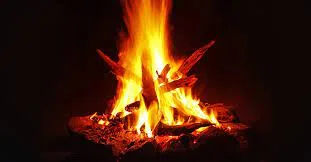 A bright campfire of stacked logs burning with tall orange and yellow flames against a dark background.