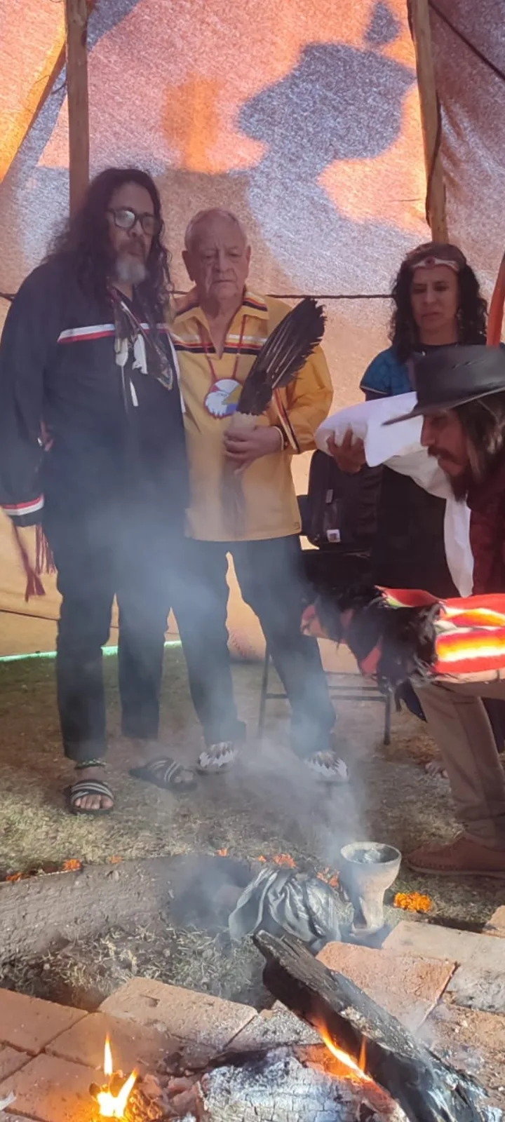 Inside a ceremonial tent, an elder in a yellow ribbon shirt holding an eagle feather fan stands with a long-haired man in black as another man bends over a smoking fire pit.